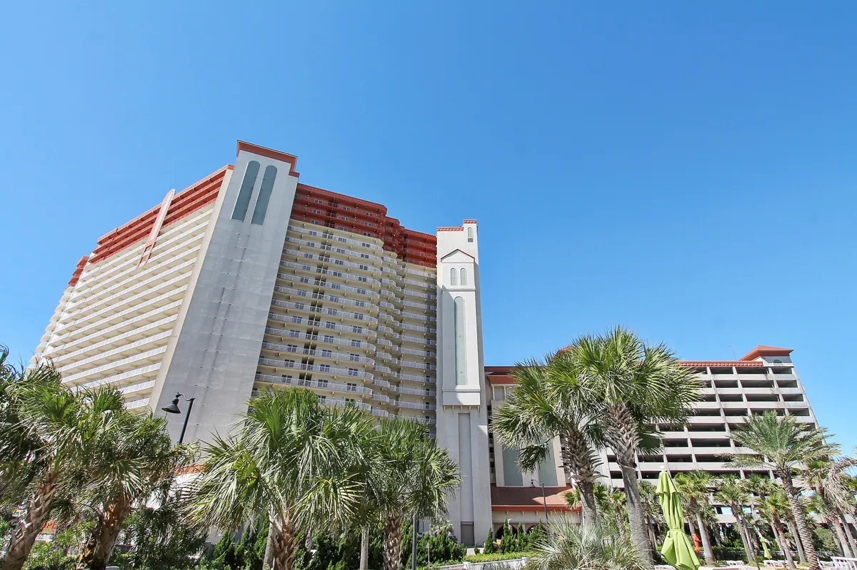A large, modern resort building framed by swaying palm trees and set against a clear blue sky. The prominent balconies suggest panoramic views of the Gulf, and the inviting exterior hints at vibrant social possibilities within and nearby. The building's proximity to the beach and amenities is visually implied.