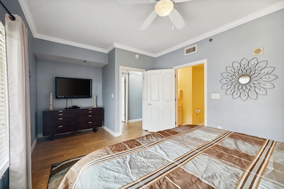 A peaceful bedroom featuring a bed with patterned bedding, a large wall mirror, and a TV placed on the dresser. The space is filled with natural light and offers a comfortable and inviting ambiance.