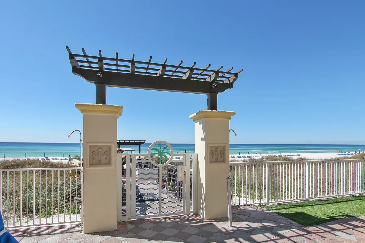 A tranquil beachside exterior featuring a pergola structure and gated entrance leading directly to the sandy shore, with sweeping views of the ocean. The setting is peaceful, highlighting the resort's private beach access and opportunities for relaxation.