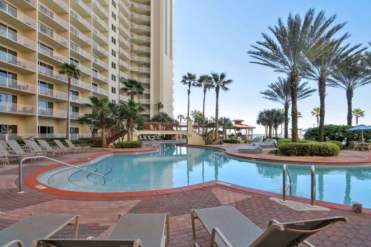 A lagoon-style swimming pool surrounded by palm trees, lounge chairs, and the balconies of a multi-story resort. The pool area is bathed in warm light, suggesting a perfect spot for relaxing, sunbathing, or enjoying evening cocktails as a group.