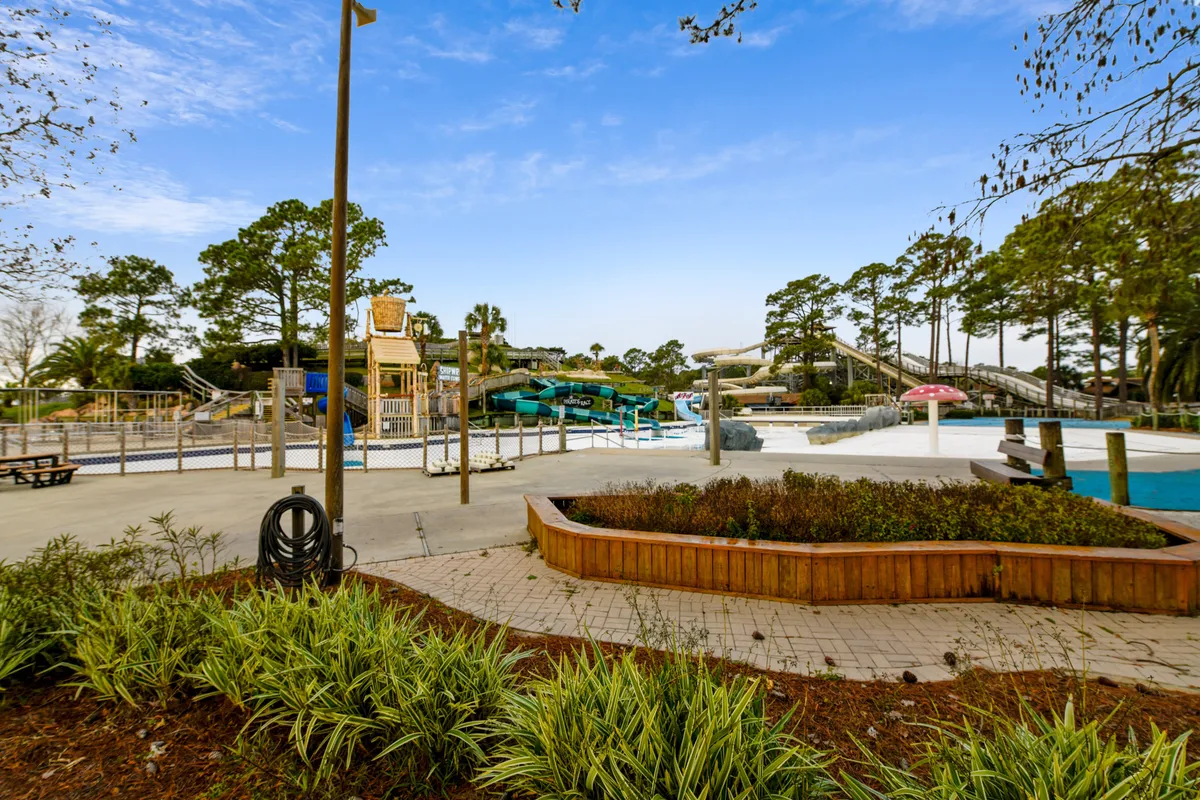 This vibrant exterior image features a lively water park area with twisting water slides, a pool, and a playground structure. Children can be seen racing down the slides while families relax and socialize around the poolside, all set against a backdrop of sunshine and cheerful colors.