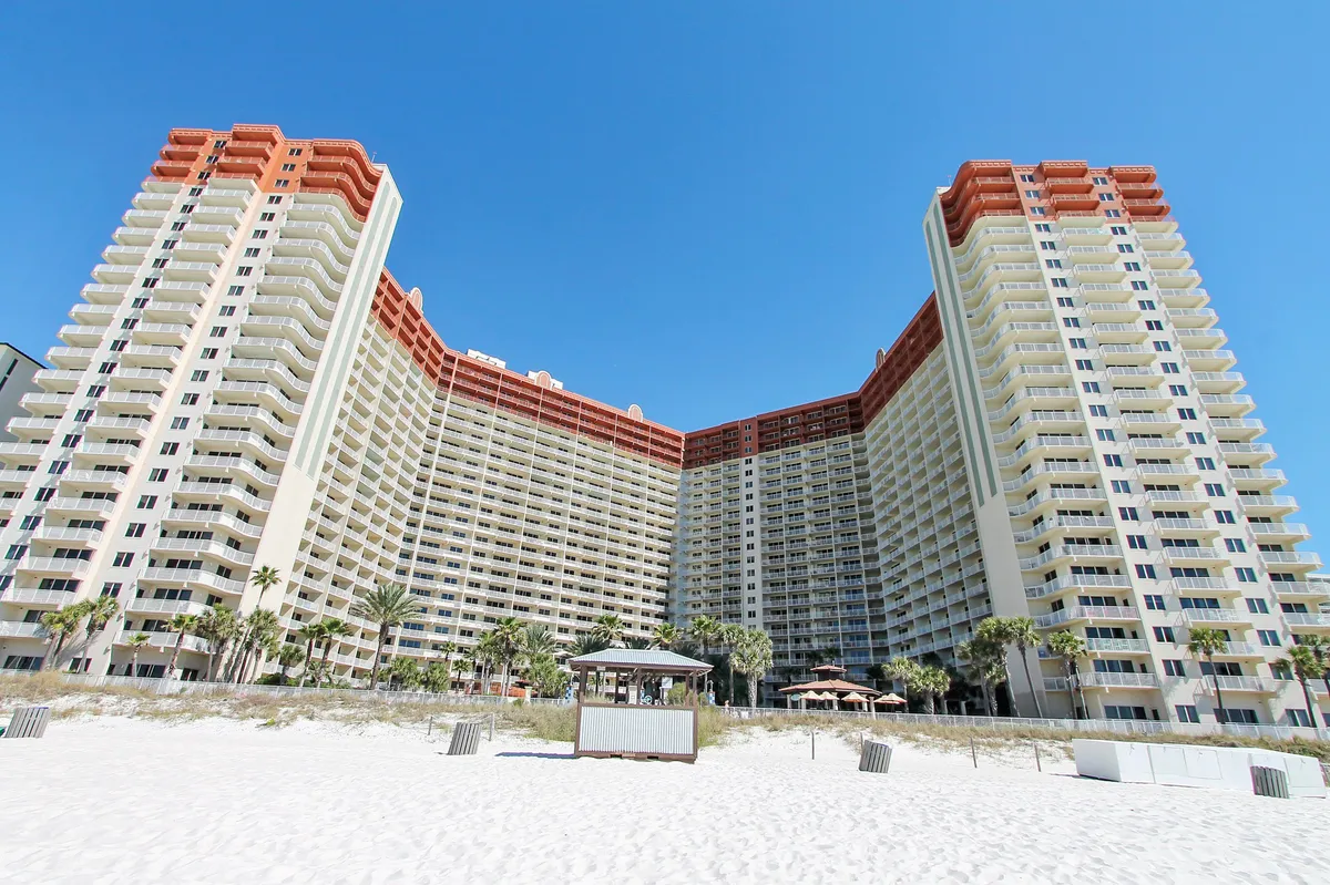 The image showcases high-rise resort buildings overlooking an expansive swimming pool area with palm trees and a gazebo along the beachfront. The pool is lively, surrounded by sunbathers and families, with a clear view to the ocean and plenty of space for poolside relaxation and games.