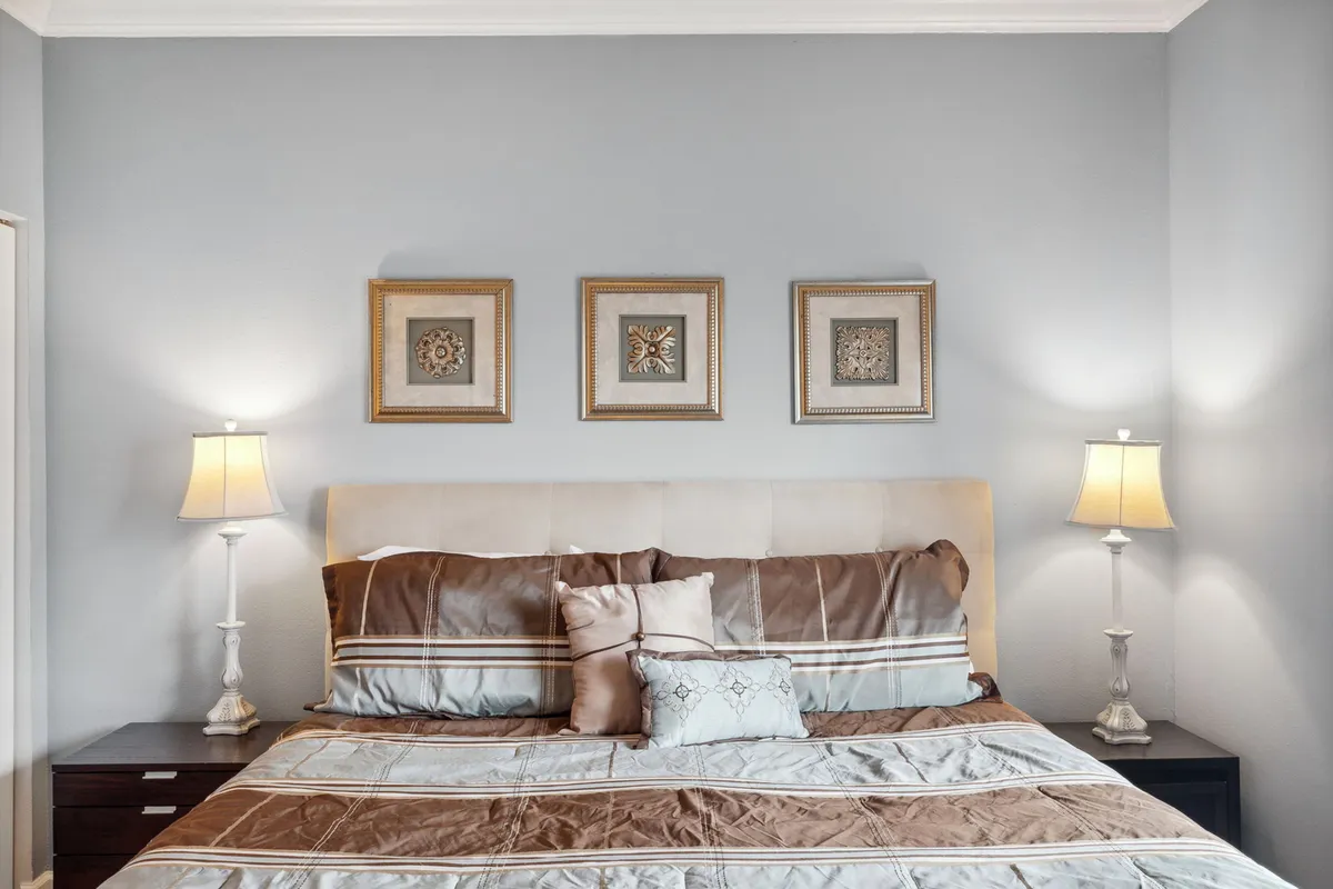 A tranquil bedroom with a bed dressed in decorative pillows, framed artwork above the headboard, and warm table lamps on either side. Soft natural light filters into the room, underscoring a sense of calm and relaxation ideal for a restful night's sleep.