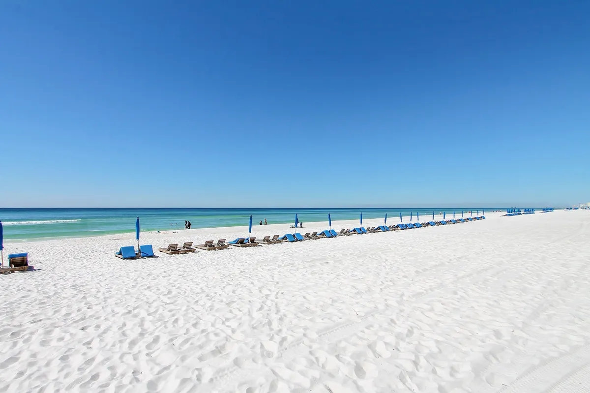 This image features inviting beach loungers and umbrellas set up on a stretch of white sand, with the blue ocean and sky in the background. It captures a tranquil coastal atmosphere, with space for sunbathing, swimming, and enjoying the shoreline.