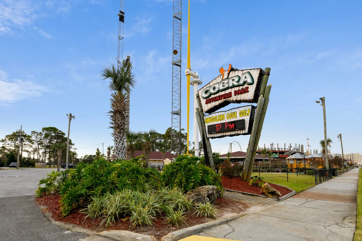 An inviting exterior shot showcases the entrance to Cobra Adventure Park, complete with a prominent sign, palm trees, and manicured landscaping. The park exudes a sense of adventure with mini-golf challenges and family-friendly attractions just beyond the entrance.