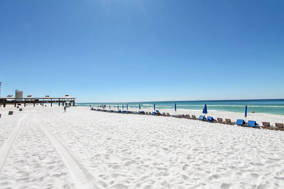 This image captures the white sandy beach lined with blue umbrellas and beach chairs, with gentle ocean waves and a pier in the background. People are enjoying activities like sunbathing, swimming, playing beach volleyball, and strolling along the shore, all under a bright, joyful sky.