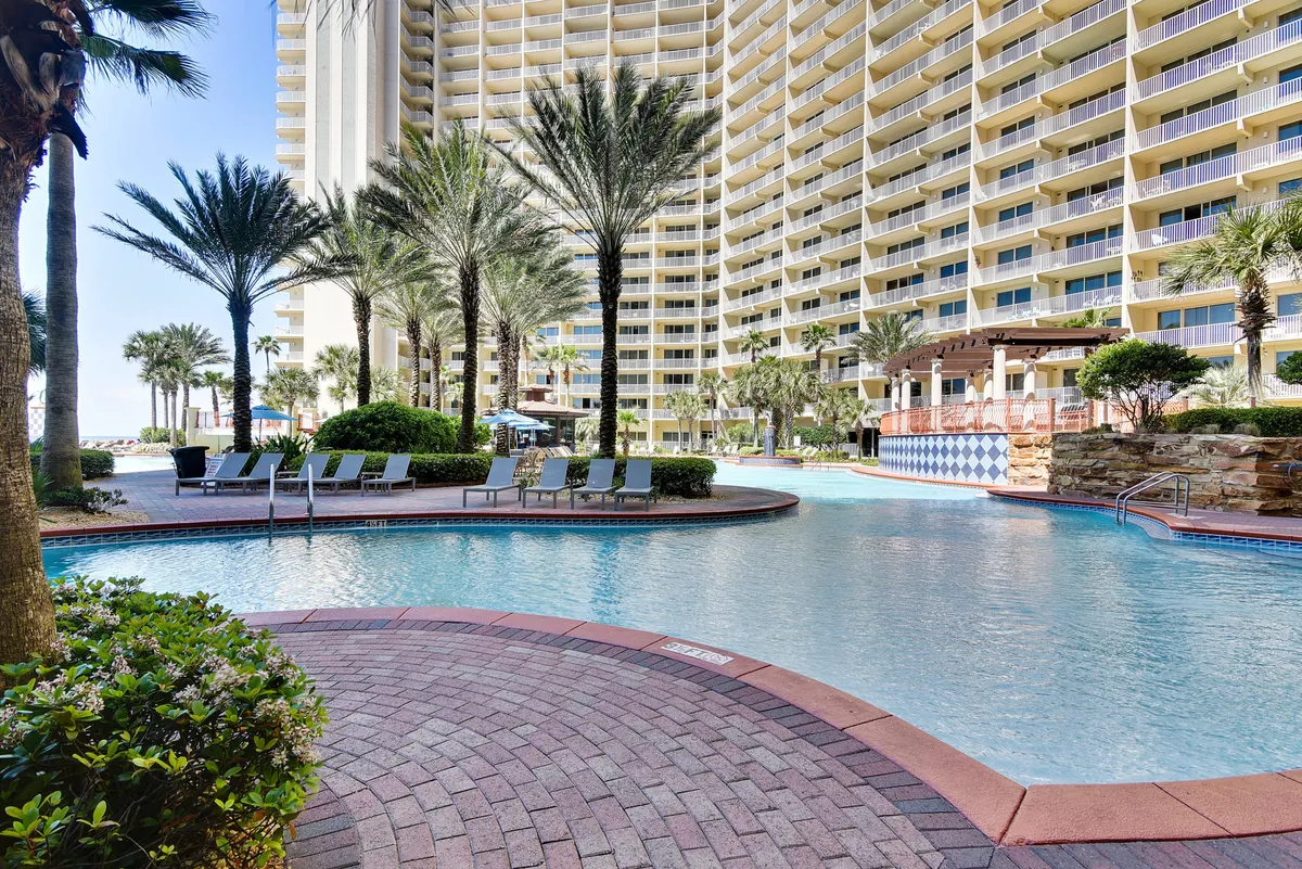 A sun-dappled swimming pool surrounded by palm trees, lounge chairs, and views of balconies above. The setting is peaceful and luxurious, suggesting relaxing swims and poolside lounging in a tropical climate.