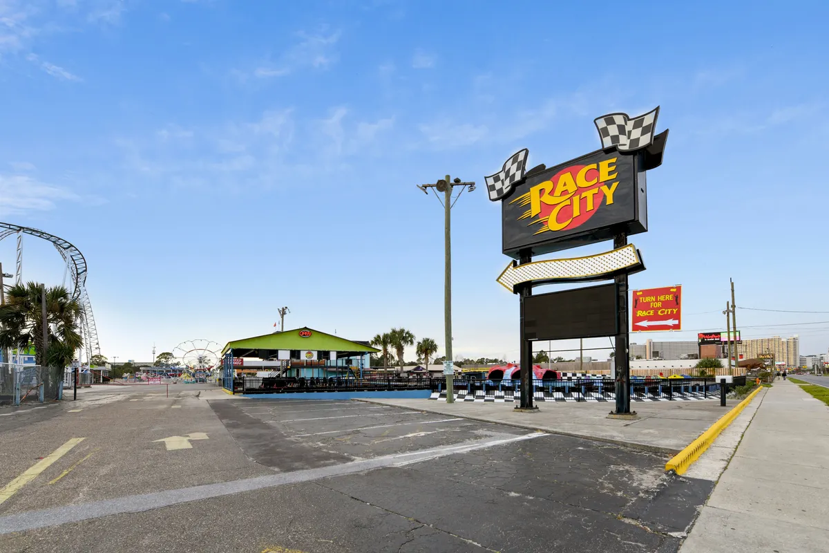 This vibrant exterior shot features the Race City amusement park sign, a go-kart track, and a ferris wheel in the background, all bathed in the golden light of late afternoon or early evening. There are families and groups enjoying amusement rides, capturing a sense of excitement and adventure.