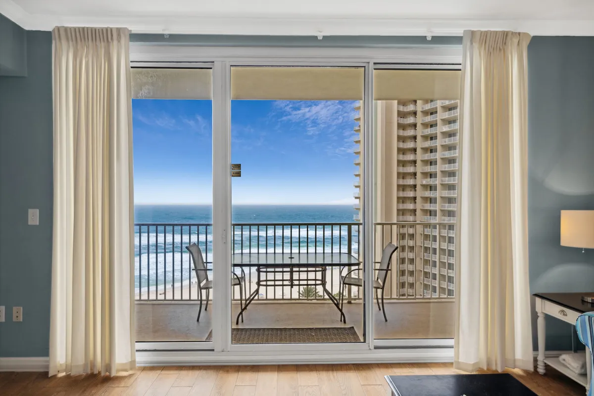 This image features floor-to-ceiling sliding glass doors opening onto a private balcony, furnished with an outdoor table and chairs. The balcony looks directly out over the Gulf, with blue sky and waves visible in the background. Soft cream curtains frame the entryway, blending the serene interior space with the breezy outdoors.