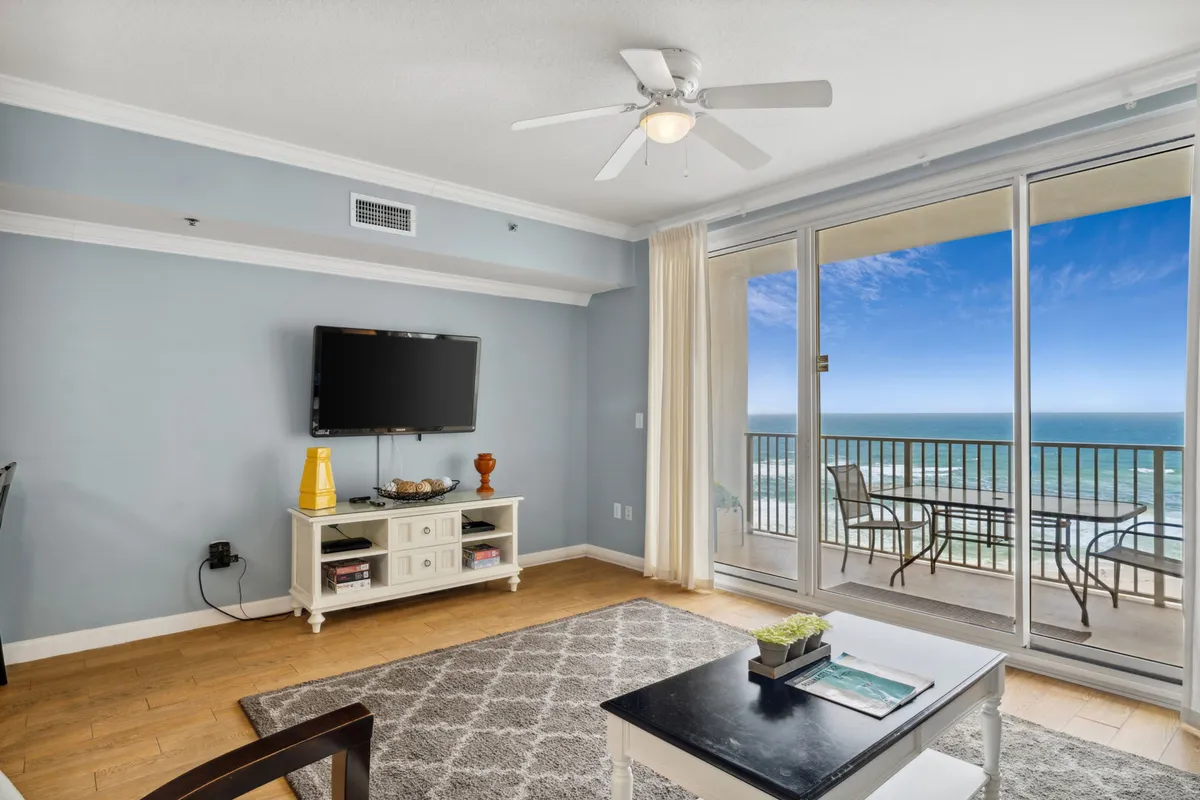 A bright, open living area with floor-to-ceiling glass sliding doors leads to a private balcony overlooking the Gulf. The room features a comfortable seating area with a flat screen TV, light wood flooring, and a patterned area rug, emphasizing natural light and a seamless indoor-outdoor connection.