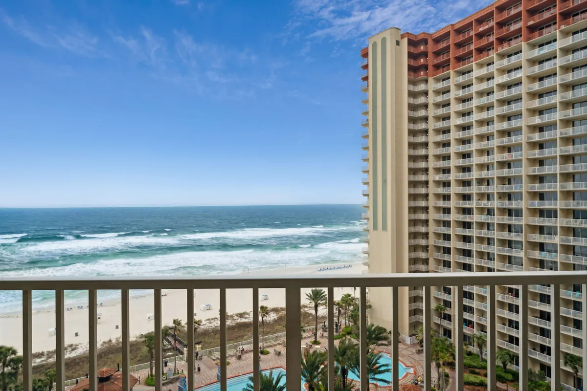 This exterior image captures the large lagoon style swimming pool with playful fountains, the high-rise condo building with balconies, and the sandy beach with the Gulf of Mexico in the background. Palm trees and a landscaped courtyard round out the inviting resort atmosphere, while families can be seen lounging by the pool and gathering in social areas.