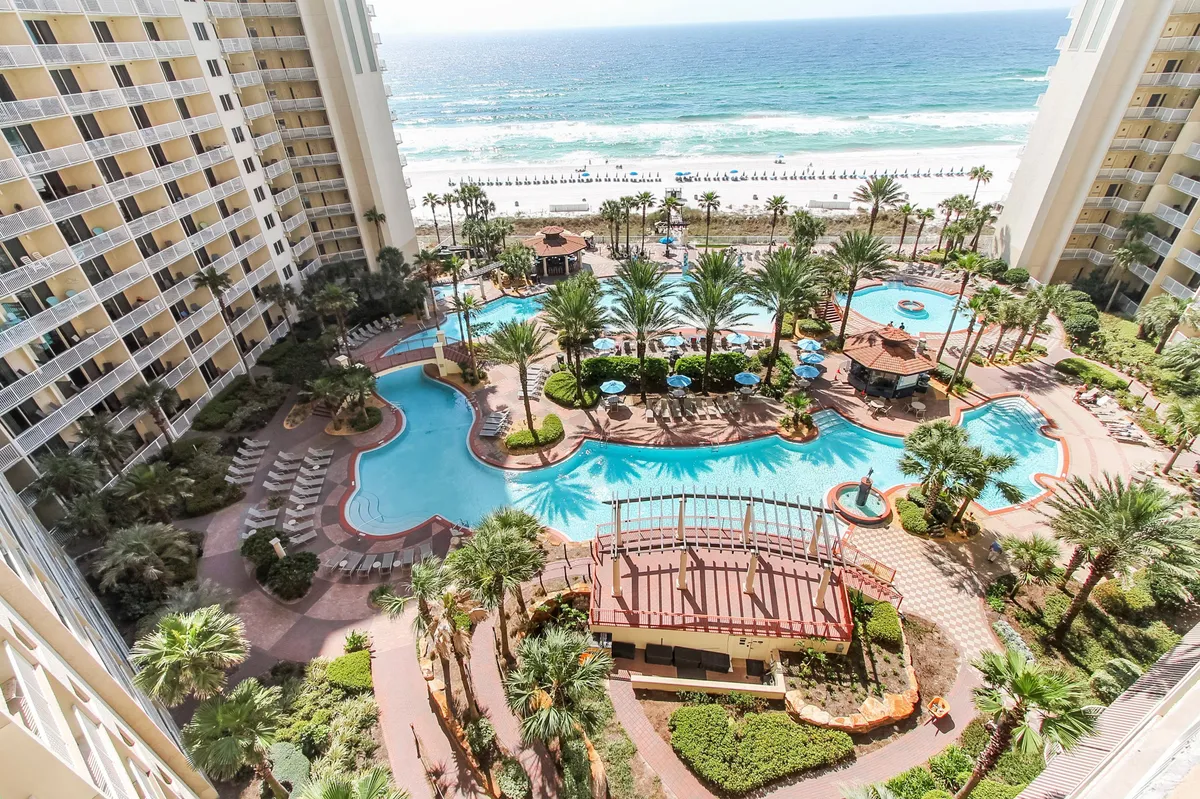 A vibrant exterior scene capturing the resort's large lagoon-style swimming pool flanked by palm trees and tropical landscaping. The poolside area features comfortable lounge chairs, tiki bars, and a view of the private beach with ocean waves rolling in the background. The multi-story resort building is visible with balconies overlooking the pool and Gulf.
