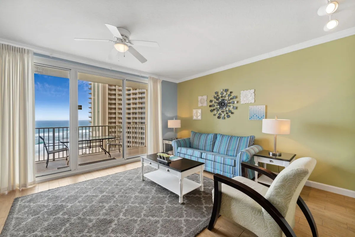 This interior scene shows a striped blue sofa facing large floor-to-ceiling sliding glass doors, which open to a furnished balcony with an ocean view. Decorative wall art and fresh natural light make the space feel calm and inviting, while the arrangement suggests easy access to the balcony for enjoying the breeze and views.