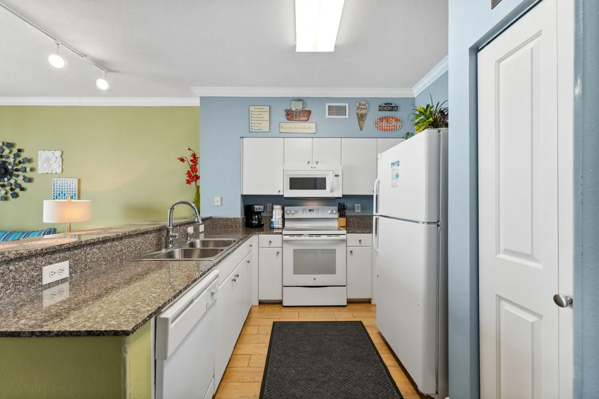 The kitchen features a granite countertop with a double sink, white appliances, and decorative wall signs above the cabinets. The open layout connects the kitchen to the living area, creating a space for meal prep, casual dining, and socializing with family or guests.