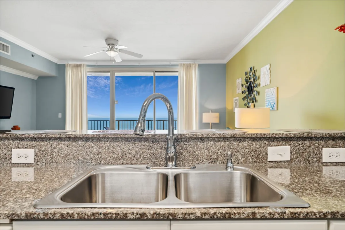 The kitchen area with a granite countertop and double sink looks out through floor-to-ceiling glass doors to a balcony, revealing a blue sky and ocean view. The open layout connects the kitchen smoothly to the living area, emphasizing the home's bright and airy design.