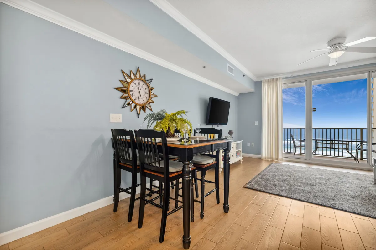 A dining area set for four is featured with a stylish decorative centerpiece. The large floor-to-ceiling windows provide expansive, unobstructed views of the Gulf, bathing the space in natural light. A wall-mounted television and decorative sunburst clock add to the inviting, sophisticated atmosphere.