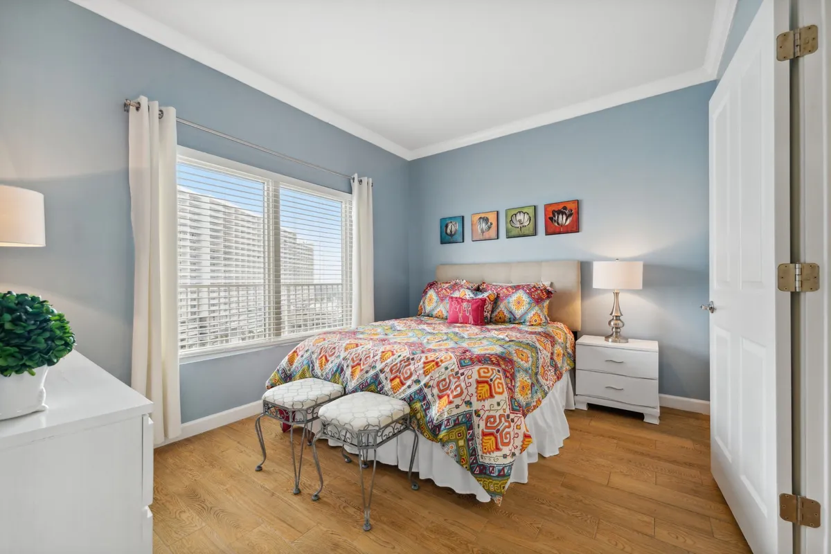 A cheerful bedroom featuring a queen-sized bed with colorful patterned bedding, two decorative stools at the foot, a nightstand with a warm-lit lamp, and a large window with white curtains allowing natural light to fill the room.
