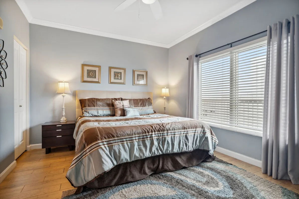 A spacious bedroom with a large bed dressed in striped bedding, warm bedside lamps on nightstands, three framed art pieces above the bed, and a large window with blinds and curtains letting in soft daylight. The mood is tranquil and inviting, suggesting a restful master suite.