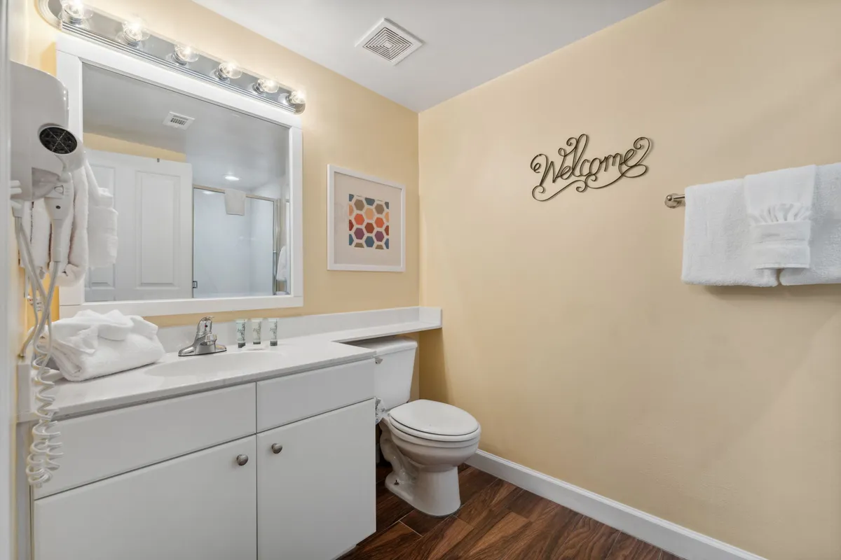 A well-lit bathroom with a large mirror and bright vanity lights, white countertop with neatly arranged toiletries, inviting 'Welcome' wall decoration, and a toilet. The space feels fresh, organized, and clean.