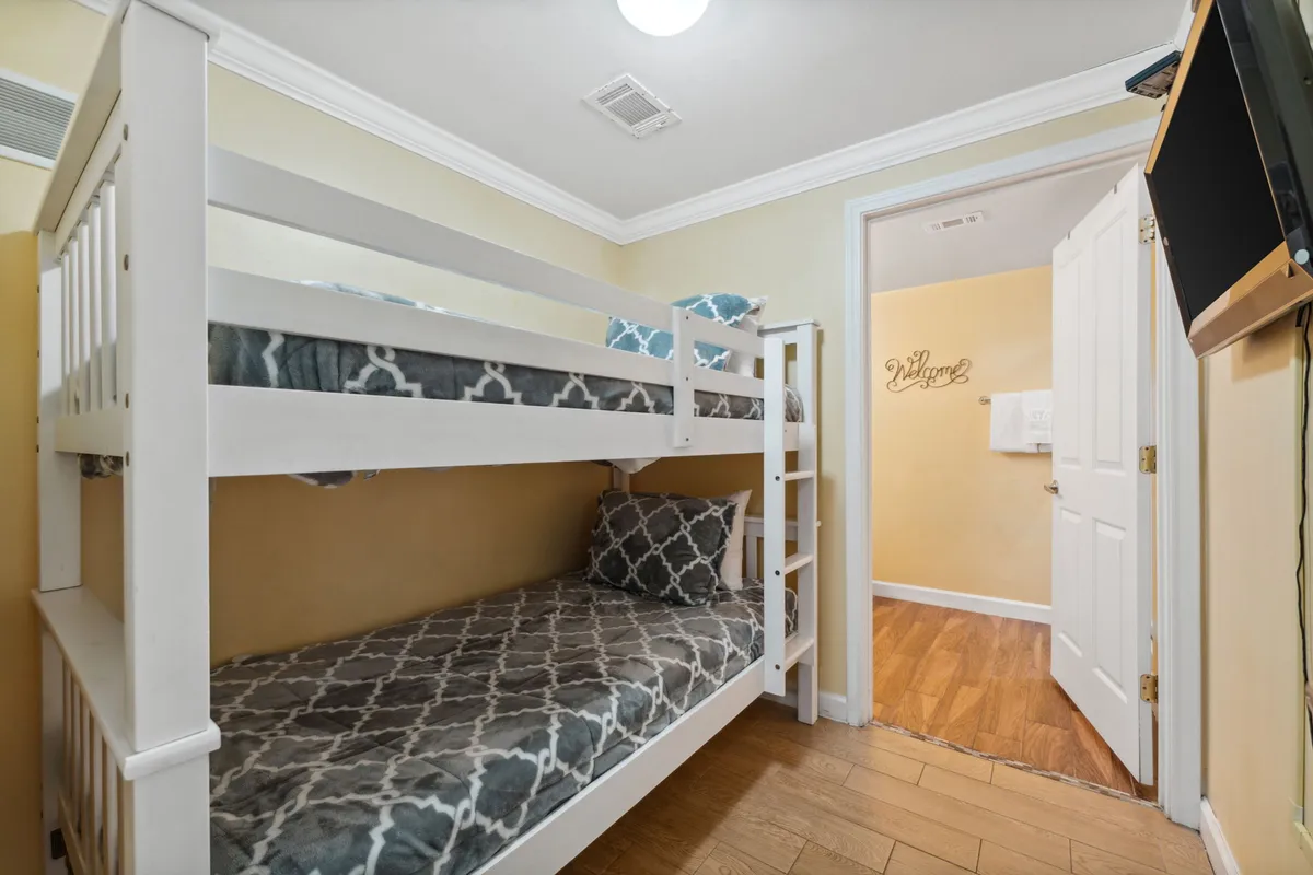 A playful bunk room with a white wooden bunk bed featuring patterned bedding, a wall-mounted flat screen TV, and an open door revealing a hallway with a 'Welcome' sign. The setting is inviting and sized for younger guests.
