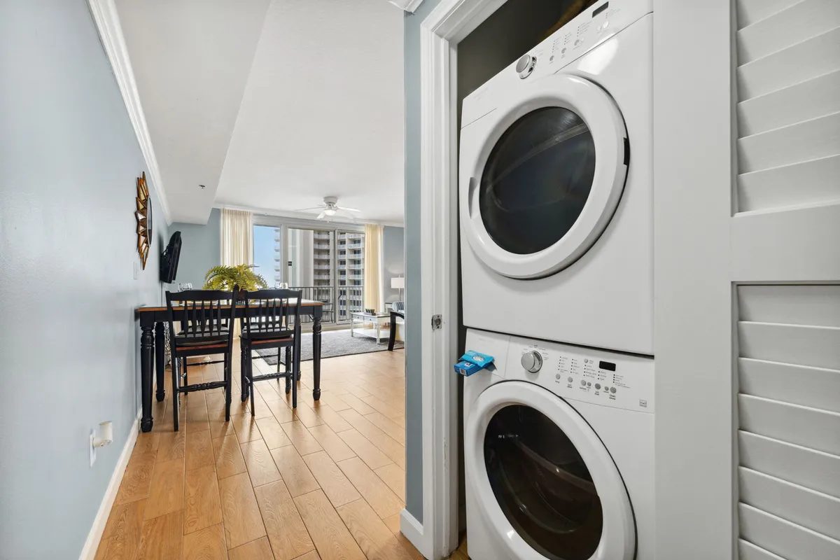 A stacked washer and dryer are tucked into a closet beside a dining table and chairs, with floor-to-ceiling windows and balcony access visible. Light wood ceramic flooring enhances the sense of space, and the open-plan layout suggests easy flow between living, dining, and kitchen spaces.