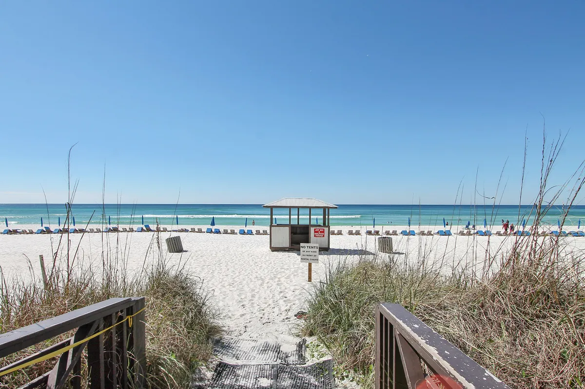 A tranquil scene of a white sandy beach lined with blue beach chairs and umbrellas, with a small lifeguard hut and the calm turquoise ocean stretching to the horizon. The setting is peaceful and inviting, perfect for morning strolls or leisurely afternoons.