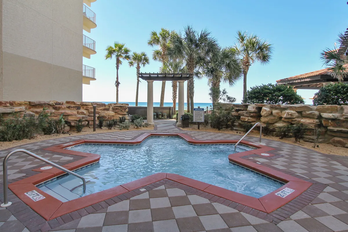 An inviting exterior featuring a star-shaped hot tub surrounded by rock wall landscaping and tall palm trees, with a pergola leading towards the sandy beach. The hot tub area looks toward the Gulf, ideal for soaking in sunset views or unwinding after an adventurous day.