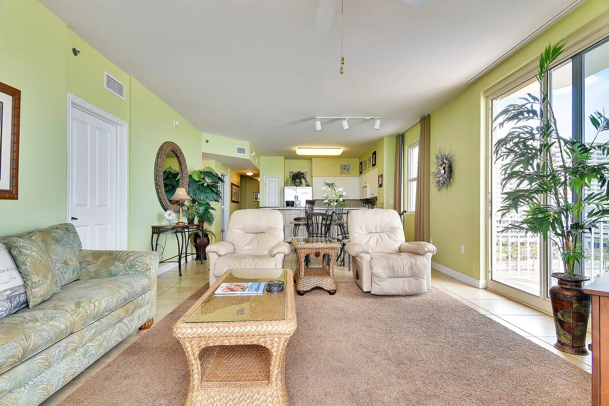 A cozy living area is depicted with two cream leather recliners, a glass coffee table with a wicker frame, large sliding glass doors leading to a balcony, and a dining area with a dark wood table and chairs. The room feels airy and welcoming, perfect for relaxing, socializing, or enjoying entertainment on the smart TV.
