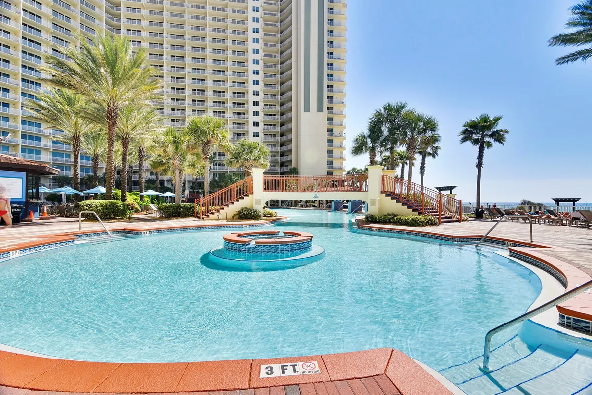 This image showcases a large lagoon-style pool with a central circular feature, surrounded by palm trees, a tall resort building with balconies, and a wooden bridge over the pool. Guests are seen lounging poolside and socializing in shaded areas, evoking year-round water fun and relaxation.