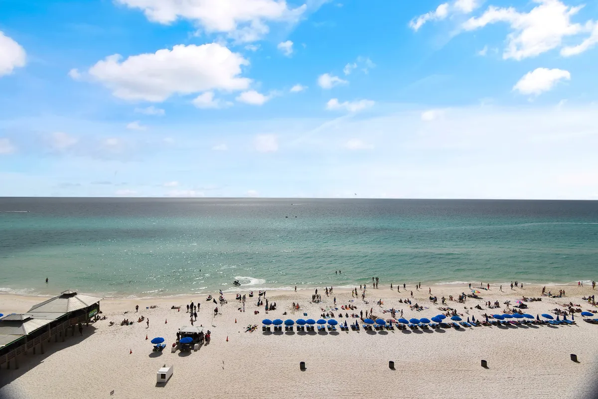 This exterior image captures a lively stretch of white sandy beach lined with blue umbrellas and people enjoying sunbathing, playing in the ocean, and socializing near a small pavilion. The turquoise waters and energetic atmosphere highlight the direct beach access and family-friendly fun described in the section.