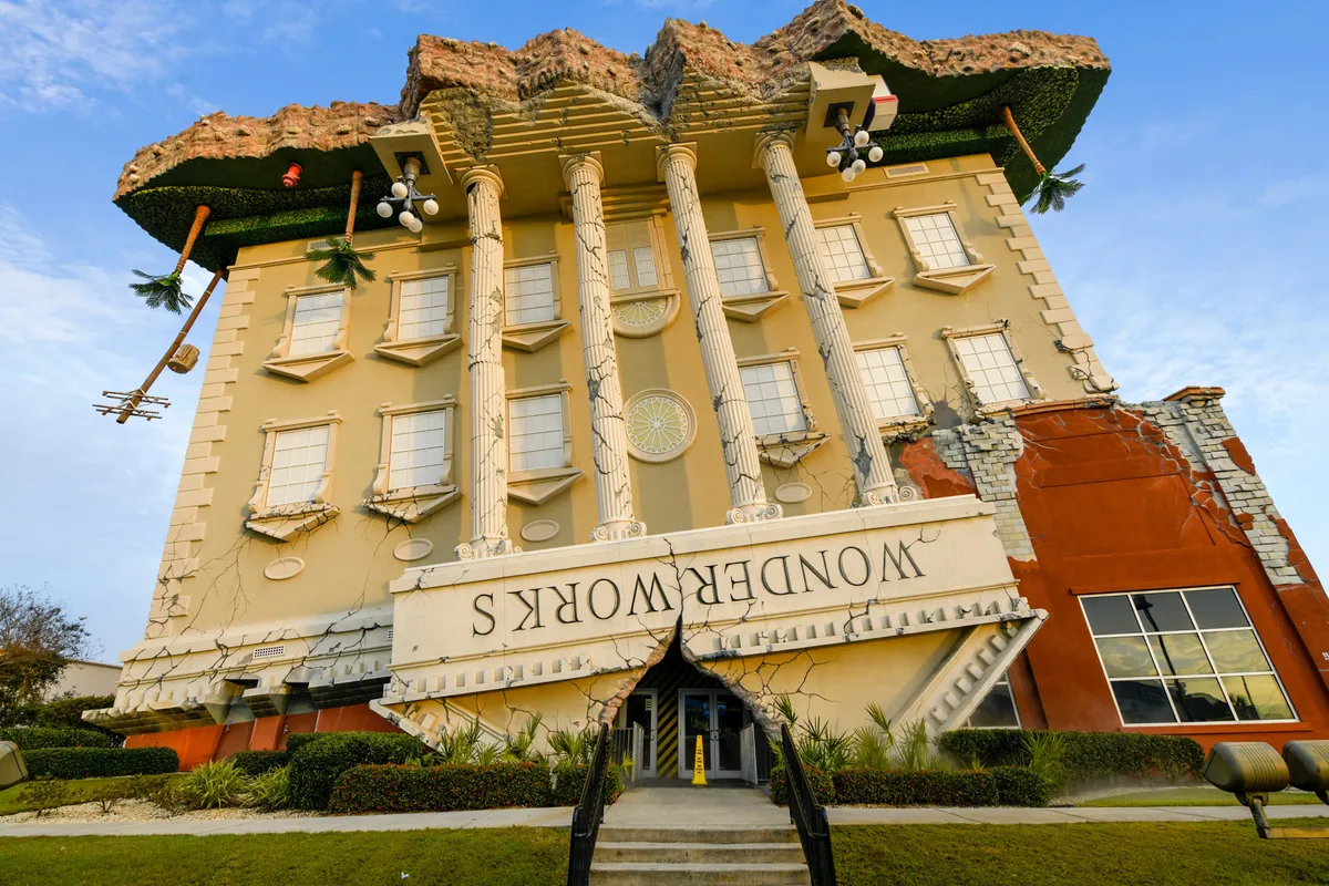 A whimsical, upside-down building with classical columns and a large 'WONDERWORKS' sign, surrounded by palm trees and a landscaped parking area. The architecture is playful and unique, suggesting fun local attractions nearby.