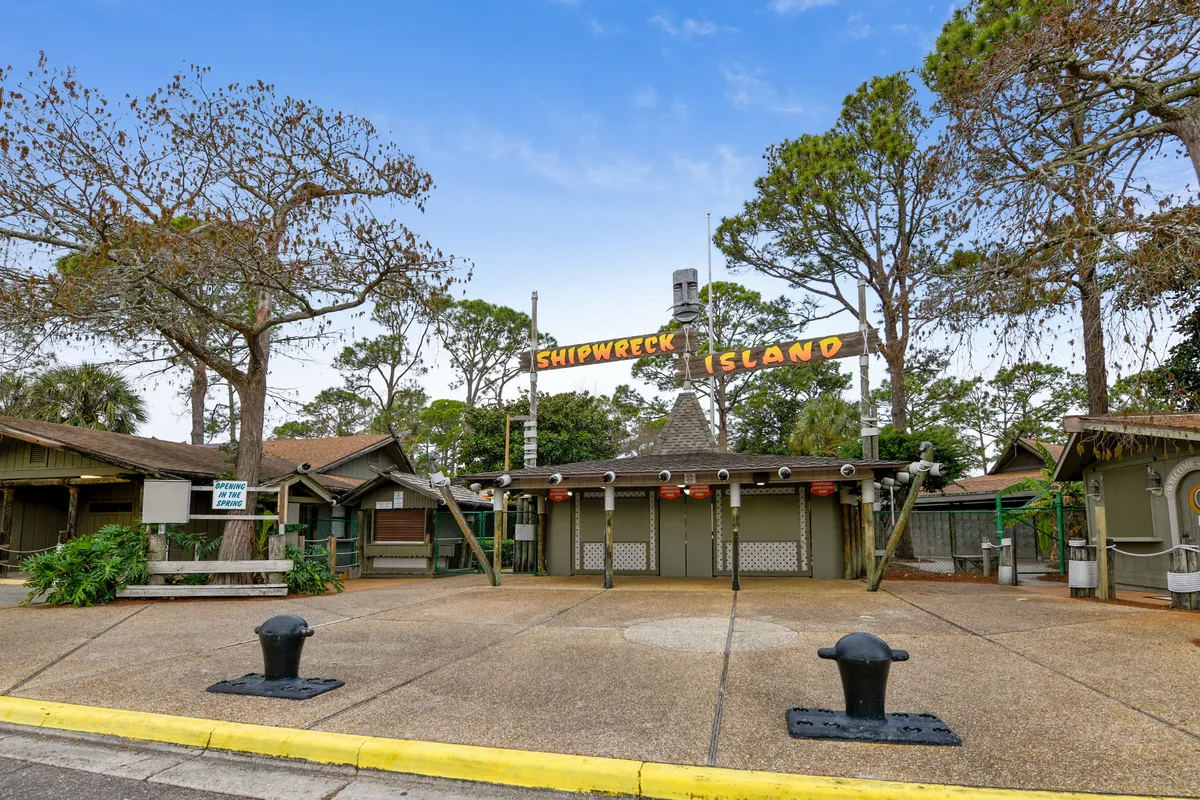 The image features the entrance to Shipwreck Island Water Park, marked by a nautical-themed wooden sign and ticket booths, with families gathering and children excited for the adventure ahead. Lush greenery surrounds the scene, signaling a day of water-based excitement and local exploration.